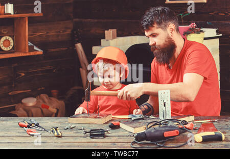 La paternità del concetto. Il ragazzo, bambino impegnato nel casco protettivo di imparare a utilizzare un martello con il papà. Padre con la barba insegnamento piccolo figlio di utilizzare strumenti, martellare, workshop su sfondo. Foto Stock