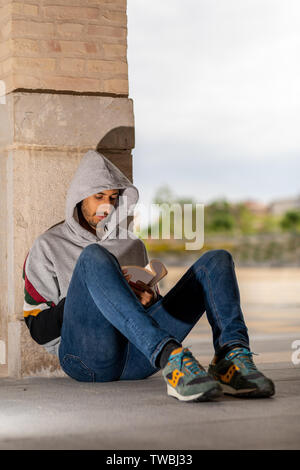 Bel giovane uomo barbuto leggendo un libro in un parco durante la mattina, indossare una felpa con cappuccio e jeans Foto Stock
