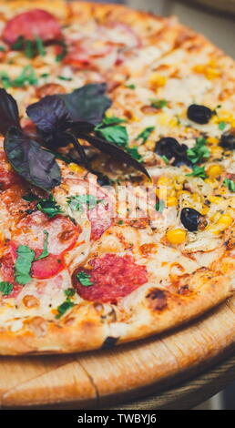 Una deliziosa pizza italiana con prosciutto di pollo, calli e olive nere su sottile sfoglia di pasta alla scrivania in legno, panorama verticale Foto Stock