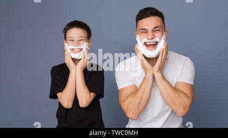 La rasatura è divertente! Papà insegnare il suo figlio per la rasatura, mettendo di schiuma sul viso, grigio Sfondo panorama Foto Stock