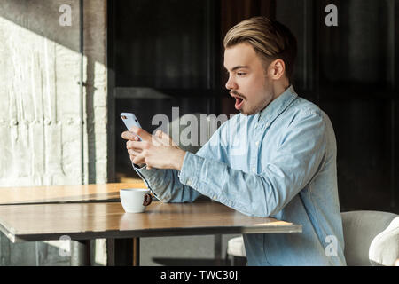 Vista laterale ritratto di scioccato giovane uomo in denim maglietta blu in seduta cafe e la lettura di notizie incredibili sul telefono con sorpresa il viso e aperto Foto Stock