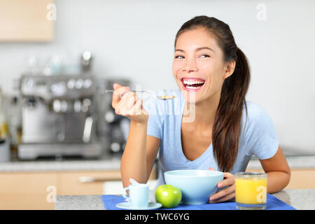 Donna di mangiare i cereali per la colazione al mattino tavola felice sorridente e mattina fresca. Giovane e bella multirazziale donna seduta nella sua cucina a casa. Razza mista Asian Caucasian modello femminile. Foto Stock