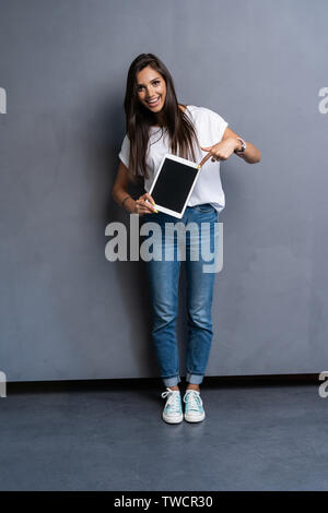 A piena lunghezza ritratto di donna graziosa azienda blank computer tavoletta su sfondo grigio Foto Stock