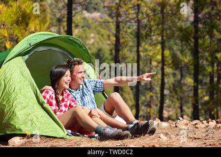 Camping matura in tenda seduto guardando a vista nella foresta. Camper sorridendo felice all'aperto nella foresta rivolta verso l. Multirazziale giovane avendo divertimento rilassante dopo l'attività all'aperto. Donna asiatica, uomo caucasico. Foto Stock