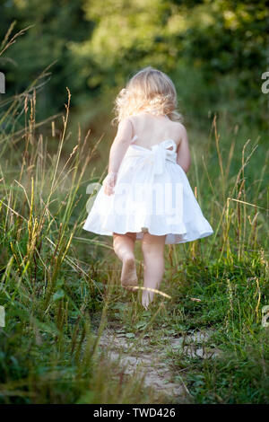 Bambina in abito bianco a piedi fino alla strada tra erba verde vista posteriore Foto Stock