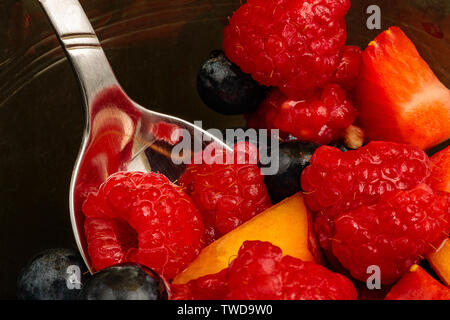 Nei pressi di macro di una ciotola di vetro di frutta con un cucchiaio e vari pezzi di frutta. Il cucchiaio è nella ciotola e riempito con i lamponi sono i principali Foto Stock