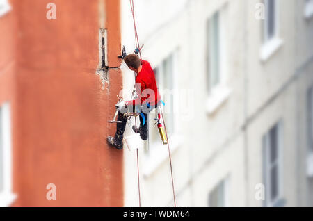 Lavoratore scalatore appeso su funi su High-Rise sfocata facciata di edificio, giunti di pulizia con ascia martello prima della sigillatura. Manutenzione dei giunti tra Pr Foto Stock