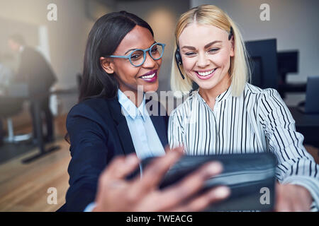 Due sorridenti giovani imprenditrici in piedi in un luminoso ufficio moderno e prendendo un selfie insieme Foto Stock