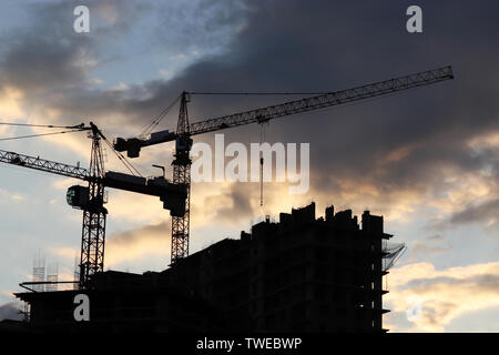 Sagome di due gru edili e gli edifici residenziali contro il cielo di sera con nuvole. Sito in costruzione sul tramonto Foto Stock