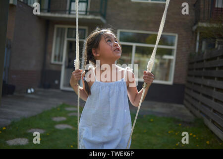 Carino giovane ragazza asiatica che indossa un abito giocando su un altalena nel giardino sul retro in un sole luminoso Foto Stock