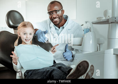 Mostrando la loro felicità. African American dentista in Grande Uniforme seduto dietro il suo piccolo paziente e godendo il suo lavoro Foto Stock