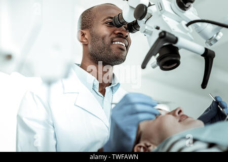 Esplorazione di equipaggiamento speciale. Bello dentista positivo cercando nel microscopio gigante durante la sessione di grave con il suo paziente Foto Stock