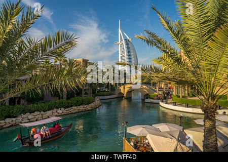 Un abra barca scivola attraverso le acque blu del Souk Madinat Jumeirah come la vela-come il Burj al Arab sorge overhead. Foto Stock