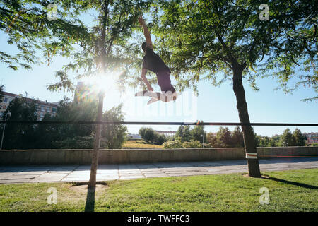 Metà adulto uomo caucasico jumping e camminare sulla corda appesa su alberi, facendo un lavoro trickline fuori di routine in Madrid, Spagna. Foto Stock
