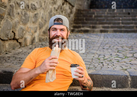 Che cosa uno snack. Uomo Barbuto mangiare malsano di hot dog snack a sandwich. Tanga e di appoggio avente hotdog snack su scale all'aperto. Ragazzo caucasico tourist gustando uno spuntino cibo sulla strada. Foto Stock