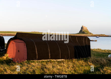 Una vecchia barca da pesca capovolta e utilizzata come capanna di deposito sulla spiaggia di Lindisfarne Holy Island con il castello di Lindisfarne Foto Stock