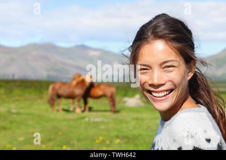 Donna su Islanda sorridente con cavalli islandesi. Ritratto di felice ragazza multiculturale che indossa un maglione islandese all'aperto permanente nel campo della natura nella parte anteriore del cavallo. Foto Stock