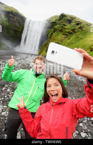 Selfie giovane prendendo selfie divertenti immagini dello smartphone di cascata all'aperto di fronte Skogafoss in Islanda. Paio di visitare famose attrazioni turistiche e punti di interesse in natura Islandese paesaggio. Foto Stock