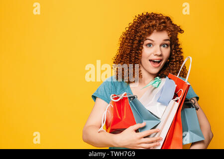 Capelli ricci donna con sorpresa faccia tenendo molte borse per lo shopping su sfondo giallo Foto Stock