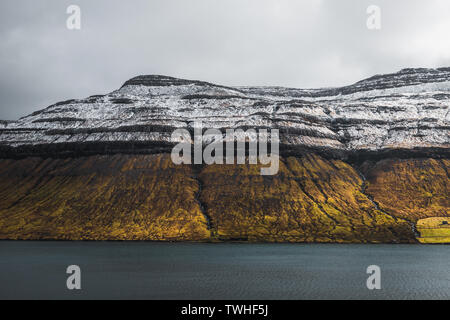 Vista sul tipico paesaggio delle isole Faeroeer con coperte di neve montagna con vasto prato arancione terra di fronte all'Oceano Atlantico (Isole Faerøer, Danimarca) Foto Stock