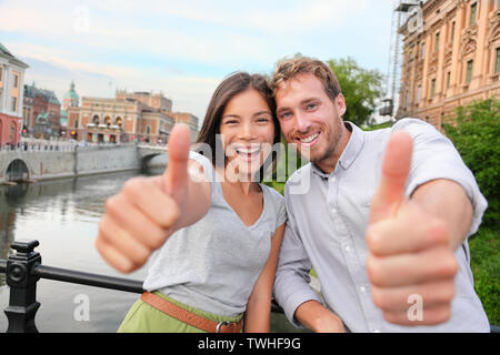 Pollice su coppia felice a Stoccolma, Svezia. La gente entusiasta di dare Thumbs up gesto guardando la fotocamera. Multirazziale coppia giovane a piedi al di fuori di Stoccolma. Uomo scandinavo, donna asiatica. Foto Stock