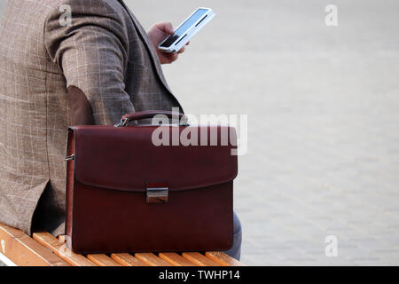 Uomo in un business suit seduta con una cartella in pelle su un banco e utilizza lo smartphone. Nozione di imprenditore, gazzetta, dipendente del governo Foto Stock