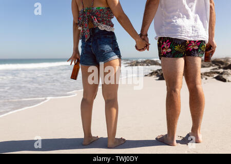 Coppia con bottiglia di birra tenendo le mani sulla spiaggia sotto il sole Foto Stock