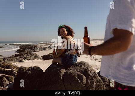 Donna con bottiglia di birra seduti sulla roccia e guardando l uomo sulla spiaggia sotto il sole Foto Stock