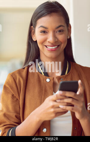 Imprenditrice guardando la fotocamera mentre si utilizza il telefono cellulare in un ufficio moderno Foto Stock