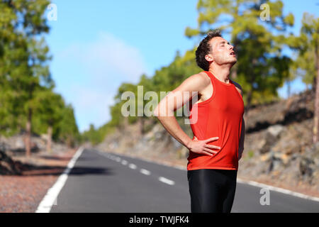 Runner in appoggio stanco ed esausto dopo l'esecuzione. Jogging uomo prendendo una pausa durante il corso di formazione esterno in sulla strada di montagna. Giovane maschio caucasico modello fitness dopo il lavoro fuori. Foto Stock
