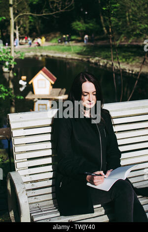 Pale scuoiati brunette lavorando sui suoi compiti (saggio o la ricerca di carta) e scrivere i suoi pensieri in un notebook. Il lago e la casa in legno Foto Stock