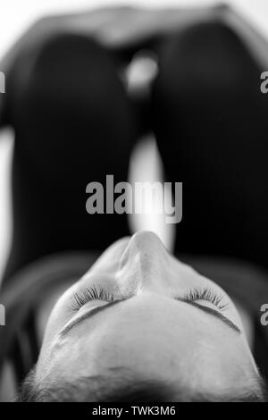 Un dark-haired brunette donna europea si trova sulla sua schiena facendo le ginocchia al petto porre o Apanasana, in bianco e nero verticale di close-up low-level vista. Foto Stock