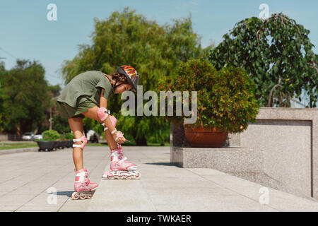 Ragazza adolescente in un casco impara a guidare su pattini a rotelle tenendo un equilibrio o roller e spin presso la città della strada nella soleggiata giornata estiva. Uno stile di vita sano, infanzia, hobby, attività per il tempo libero. Foto Stock