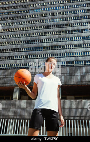 Ritratto di una giovane donna giocatore di basket tenendo la palla contro un pesante edificio urbano Foto Stock