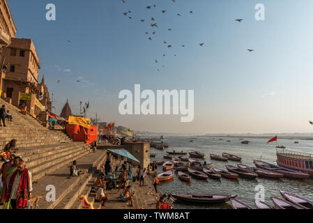 Varanasi Ghat su un morbido inverno mattina di sole con le barche e i pellegrini che si muove intorno al ghats. Uccelli al mattino sono battenti nel profondo blu skyof Varanasi Foto Stock