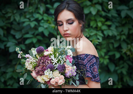 Ritratto di una giovane e bella donna con perfetto Trucco e acconciatura tenendo un mazzo di fiori selvatici. In piedi contro la parete di edera. Conce Foto Stock