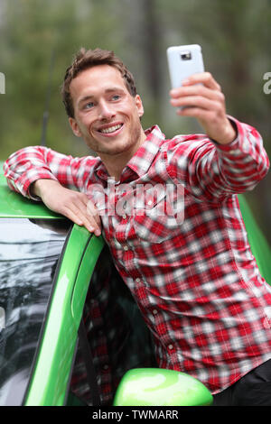 Autista con Auto prendendo selfie foto con lo smartphone dopo la guida nel nuovo verde auto. Uomo felice di scattare una foto con la fotocamera dello smartphone durante il viaggio viaggio su strada. Giovani caucasici modello maschio nel suo 20s. Foto Stock