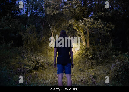 Vista posteriore del giovane uomo che indossa pantaloni corti e t-shirt e foresta di illuminazione con lampada da testa durante il periodo estivo Foto Stock