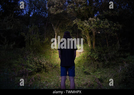 Vista posteriore del giovane uomo che indossa pantaloni corti e t-shirt e foresta di illuminazione con lampada da testa durante il periodo estivo Foto Stock