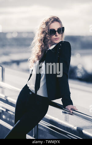 Moda giovane bionda business donna appoggiata sulla ringhiera elegante modello femminile indossando giacca nera e occhiali da sole Foto Stock