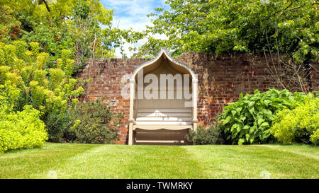 Vintage appartata panca in legno da un muro di mattoni, in un elegante giardino paesaggistico, in una giornata di sole in un paesaggio inglese . Foto Stock