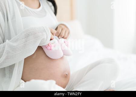 Donna incinta con scarpe per bambini sul letto di casa. Godendo del suo periodo speciale. Foto Stock