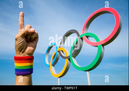 Londra - 4 Maggio 2019: Una mano che indossa il Gay Pride arcobaleno braccialetto colorato di punti al cielo con un numero di un gesto di fronte anelli olimpici. Foto Stock