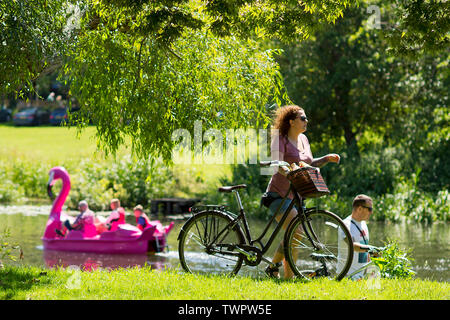 Una donna che spinge la sua bicicletta lungo la riva del fiume come persone su un pedalò godetevi il meteo sul fiume Avon in Warwick durante una calda giornata d'estate. 22.06.2019 Foto Stock