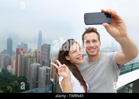 Hong Kong Victoria Peak turisti giovane tenendo selfie foto con lo smartphone gode di vista su Hong Kong e del porto di Victoria. Giovani multietnica felice coppia che viaggia in Asia. Foto Stock