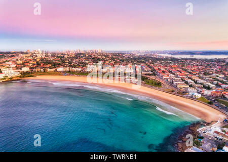 Pulire arco di larghe spiagge sabbiose di Bondi Beach a Sydney all'alba con cielo rosa e calma onde lisce floating surfers e circondata da sobborghi orientali. Foto Stock