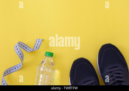 Fitness laici piana. Uno stile di vita sano e sport concept. Sneakers nero, metro a nastro e una bottiglia di acqua su uno sfondo giallo. Foto Stock