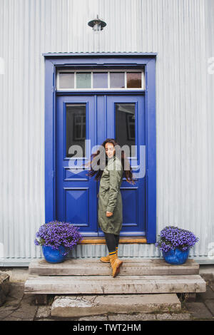 Curly brunette donna in camicia cachi e stivali arancione in piedi nella parte anteriore della porta blu e viola e vasi di fiori. Moda autunno look. Scandinavian house fa Foto Stock