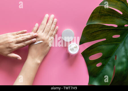 Dermocosmesi naturale concetto. donna applicare la crema bianca su le mani su sfondo rosa con un vasetto di crema cosmetica decorare con fresco verde lasciare con copia Foto Stock