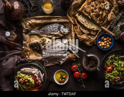 Cotta dorado pesce sulla tavola scuro con insalatiera , focaccia, pomodoro e olive, vista dall'alto. Mediterraneo il pranzo o la cena. Il cibo italiano concept Foto Stock